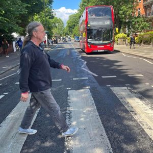 A man walks across a zebra crossing while a red double-decker bus approaches in the background on a sunny day.