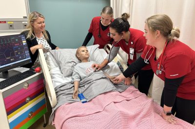 Four nursing students in red uniforms practice on a medical mannequin in a hospital bed, with a heart rate monitor nearby. An instructor observes the session.