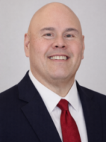 Bald man in a dark suit, white shirt, and red tie, smiling at the camera against a plain light background.